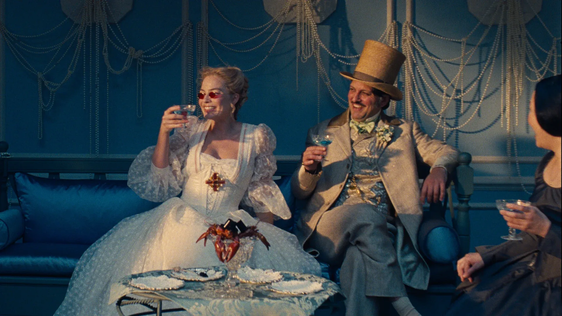 Margot Robbie and Shazad Latif, as Catherine and Edgar Linton, sipping drinks and laughing at a party, in the film Wuthering Heights.
